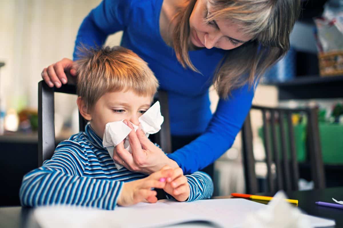 Mother leaning over child in kitchen chair as they hold a tissue up to a sitting child's nose